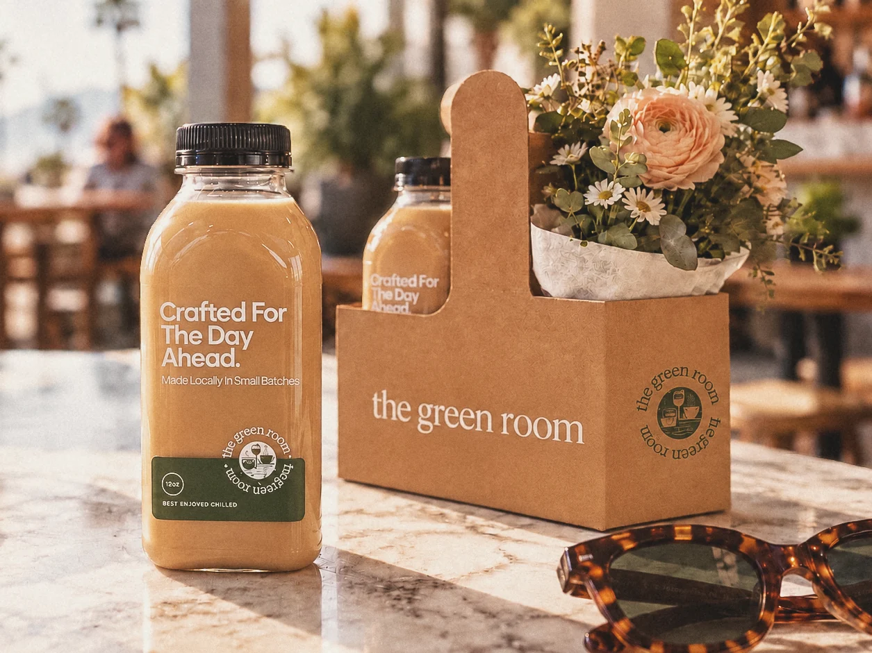 Two juice bottles and a kraft carrier on a marble cafe table, the carrier printed with the green room mark, a bunch of flowers behind it.